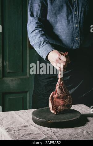 Des Menschen Hände halten Roh ungekocht Black Angus Rind tomahawk Steak auf Knochen auf Leinen Tischdecke. Im rustikalen Stil Stockfoto