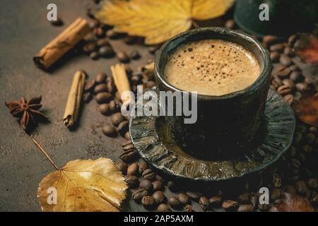 Schwarzen Kaffee Espresso mit Schaumstoff in schwarz Keramiktasse mit Untertasse, Herbstblätter, Gewürze und gerösteten Bohnen über braune Textur Hintergrund. Stockfoto