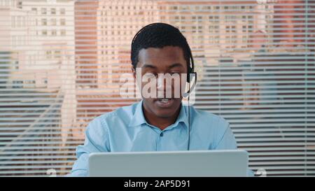Black man im Headset spricht mit jemandem und arbeitet am Computer. Er arbeitet im modernen internationalen Kundensupport. Im Hintergrund befinden sich Wolkenkratzer. Stockfoto