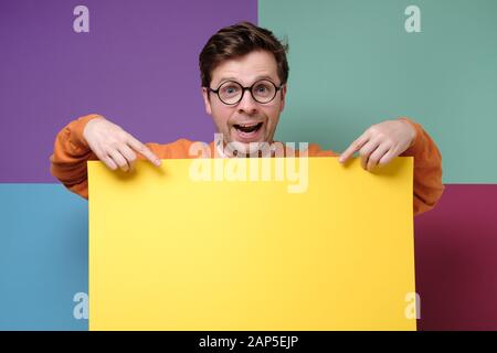 Junger positiver kaukasischer Mann, der stehend ist, hält leeres Brett, gutaussehender Kerl in einer Brille, die leeren gelben Werberaum zeigt. Studio gedreht Stockfoto