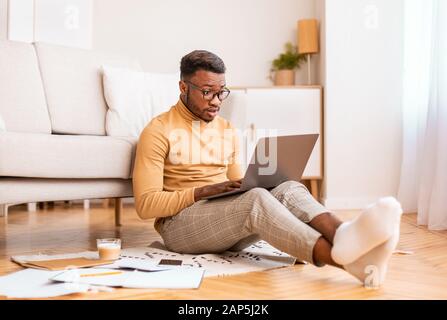 Schockierter Mann, der Nachrichten auf einem Laptop liest, der im Innenbereich auf dem Boden sitzt Stockfoto