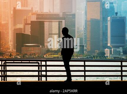 Silhouette eines Mannes auf der Suche nach Handy mit Hong Kong Island Skyline der Stadt Hintergrund Stockfoto