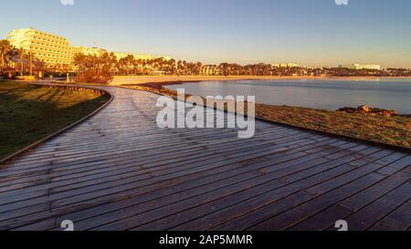 Boardwalk aus Holz bei Sonnenaufgang führt zu einem Ferienort, das von Morgensonne, Strand und Bucht mit Hotels, Restaurants, Cafés und Palmen, Sa Coma, Mallo, angezündet wird Stockfoto