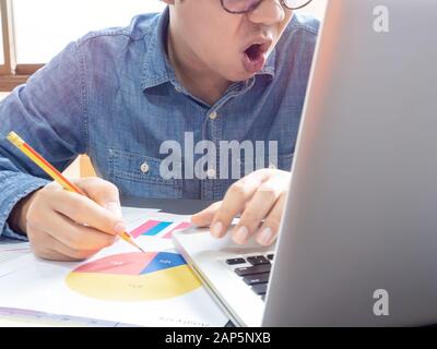 Asiatische Geschäftsleute tragen blaues Hemd und Brille mit Bleistift und fühlen sich schockiert, wenn sie mit einem Laptop auf dem Tisch in der Nähe des Fensters im Büro arbeiten. Stockfoto