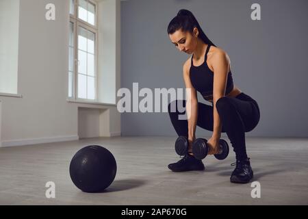 Squat-Übungen. Mädchen in schwarzer Sportkleidung mit Hanteln in den Händen, die in einem Raum hocken. Stockfoto