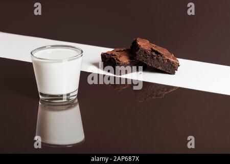 Kuschelige Schokoladen-Brownies auf weißem Hintergrund und Glas Milch auf braunem Hintergrund. Duoton-Bild von Brownies und Milch. Schokoladendessert und Milch. Stockfoto