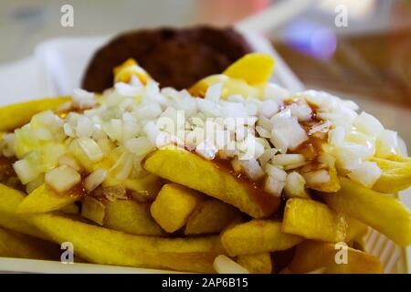 Frikandel mit Pommes Spezial: Blick auf den Haufen Pommes frites mit fetten, klebrigen Mayonnaise, Ketchup und Zwiebeln in weißer Plastikschale. Verschwommene Fleischerba Stockfoto