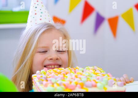 Blondes kaukasisches Mädchen, das hinter Geburtstagskuchen mit lächelndem Gesicht auf Geburtstagsfeier herausplaubert. Festlicher bunter Hintergrund mit Ballons. Stockfoto