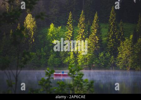 Der Mann auf dem kleinen Boot genießt den frühen Morgen mit dem Paddeln am nebligen See umgeben von Wald. Stockfoto