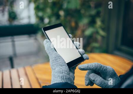 Nah-Handy mit leerem weißen Bildschirm in weiblichen Händen in Handschuhen im Freien. Stockfoto