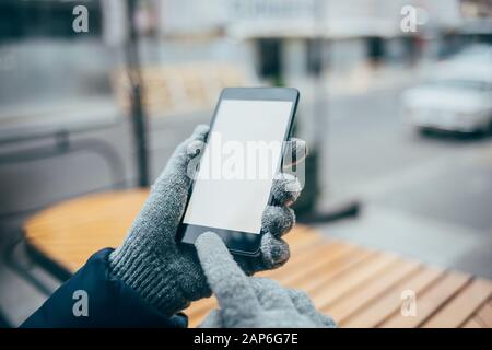 Handschuhe für Frauen mit Handy und leerem Bildschirm auf der Straße in der Stadt am Wintertag. Stockfoto