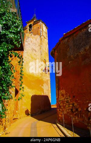 Blick nach oben über Stufen und eine schmale Gasse auf dem ockerfarbenen Kirchturm der Festung gegen den blauen Himmel - Roussillon en Provence, Frankreich Stockfoto