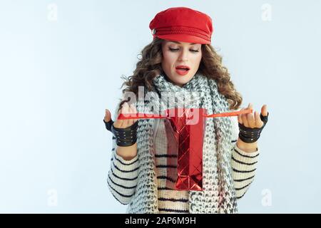 Überrascht trendy Frau mit langen brünetten Haar in Pullover, Schal und Red hat sich innerhalb der roten Einkaufstasche im Winter hellblauen Hintergrund isoliert. Stockfoto