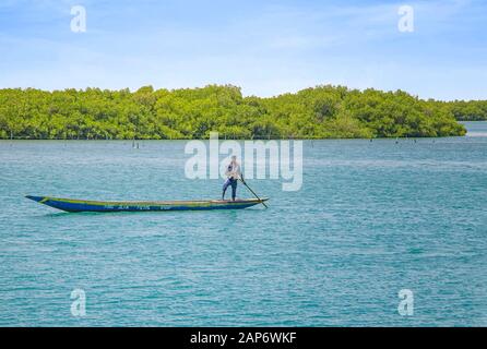 Fadiouth, Senegal, AFRIKA - 26. April 2019: Unidentifizierter senegalesischer Mann fährt mit einem typischen Holzkanu in einer See-Lagune und fischt. Hinter ihm sind es Stockfoto