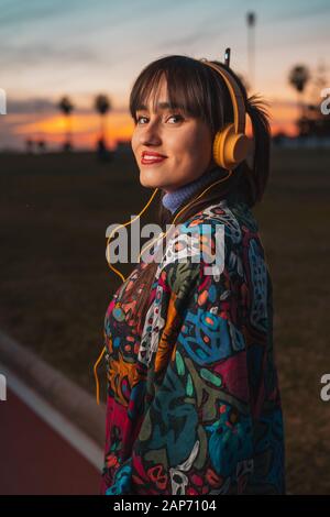 Casual schöne kaukasische braune Haarfrau Musik hören mit gelben Kopfhörern Blick auf die Kamera in einem Stadtpark während des erstaunlichen Sonnenuntergangs. Musik conce Stockfoto