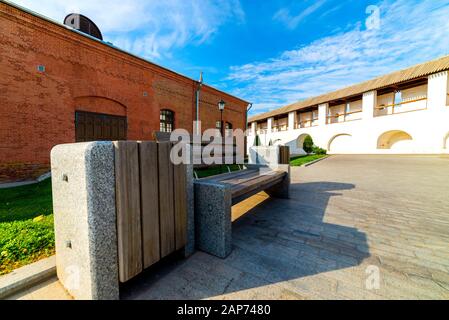 Ein mit Stein und Holzbohlen verzierter Mülleimer im Freien steht auf einem Gehweg aus grauen Fliesen in der Nähe von Holzbänken mit Blumentöpfen für Büsche. Stockfoto