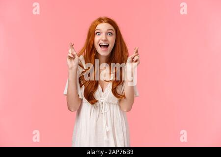 Glückliches gut aussehendes glückliches, aufgeregtes Mädchen aus der rotkopfkaukasie, das ihren Traum sah, wurde wahr, nachdem sie mit gekreuzten Fingern betete, staunte und überraschte aussehende Stockfoto