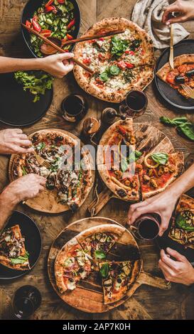 Freunde in Pizza Party Abendessen. Flachbild-lay von Menschen essen verschiedene italienische Pizza, Salat und trinken Rotwein über Holz- Tabelle, Ansicht von oben Stockfoto