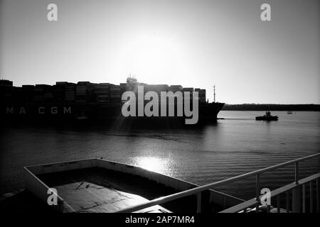 Containerschiff, das am späten Januarnachmittag in den Hafen von Southampton geführt wird, während die Sonne sich auf den Hintergrund vorbereitet Stockfoto