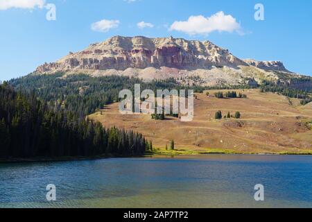 Eine bunte Felswand aus Sandstein blickt auf eine Bergwiese, einen Kiefernwald und einen schönen See. Stockfoto