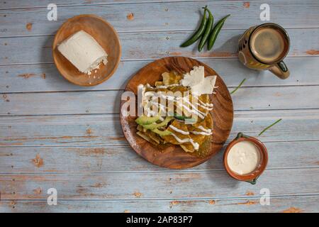 Präsentation mexikanischer Speisen mit Chilaquiles in grüner Soße, mexikanischer Kaffee in folklorischer Tasse mit Käse, Sahne und Chilipern auf blauem Holztabl Stockfoto