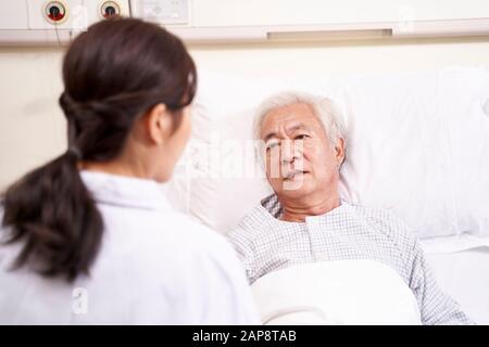 Asiatischer alter Mann, der im Bett liegt und mit junger Ärztin in der Krankenstation spricht Stockfoto