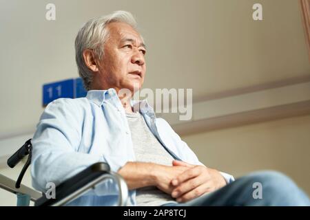 Älterer asiatischer Mann sitzt im Rollstuhl im Pflegeheim mit ernsthaftem Gesichtsausdruck Stockfoto