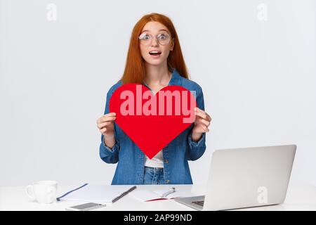 Amüsante und träumerisch überraschte Rotkopfgirl erhielt großes rotes Valentinstag Herz von einem Kollegen, der in der Nähe eines Arbeitstisches mit Geschenk stand und kgaste Stockfoto