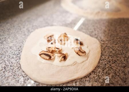 Pizza kochen. Legt Champignons und Sahnesauce aus, Zutaten auf der Teigvorform. Nahaufnahme der Küchenchef-Bäckerei in einer einheitlichen weißen Schürze in der Küche Stockfoto