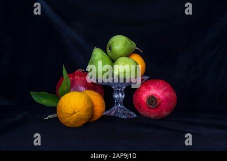 Stillleben. Birnen in einer Glasvase, Orangen und Granatäpfchen auf schwarzem Hintergrund Stockfoto