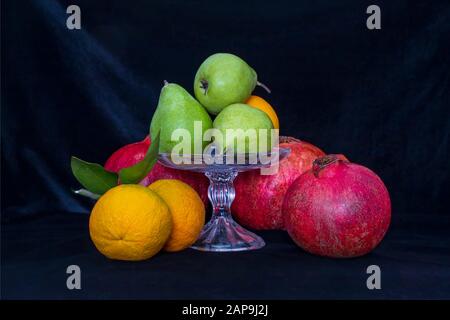 Stillleben. Birnen in einer Glasvase, Orangen und Granatäpfchen auf schwarzem Hintergrund Stockfoto