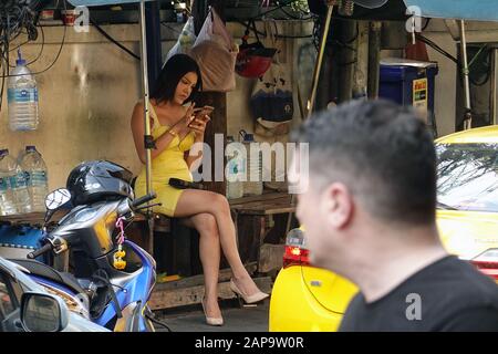 Bangkok, Thailand - 26. Dezember 2019: Transgender-Frau, Marienkind, sitzt auf der Bank und benutzt ihr Telefon. Mann im Vordergrund, der sie beobachtet. Stockfoto