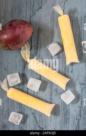 Drei cremige Mango-Lollies auf blauem Grund mit ganzen Mango-Früchten und Eiswürfeln Stockfoto