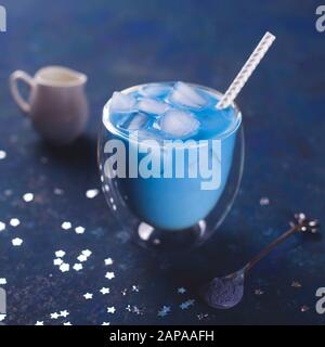 Ein Glas kaltes blau Matcha Tee mit Eis mit Strohhalm, einen Löffel Kaffee Pulver und Milchmann auf einem blauen Tisch. Farbe des Jahres 2020 Classic blau Konzept. Stockfoto