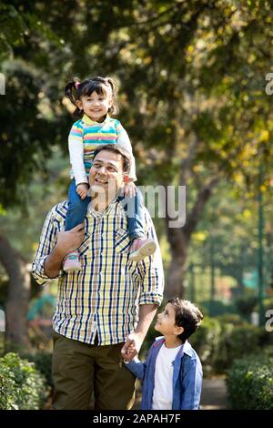 Vater, der Tochter auf den Schultern trägt und die Hände seines Sohnes im Park hält Stockfoto
