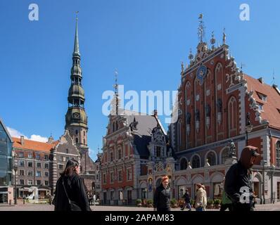 Frühlingstag blauer Himmel in der Altstadt von Riga mit Menschen, die über einen Platz, Ratslaukums, an einem sonnigen Tag mit historischen Gebäuden im Hintergrund laufen. Stockfoto