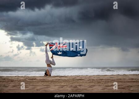 Australische Flagge, die im Wind weht und an den Füßen weiblicher Yogi hängt, die an einem australischen Strand mit dramatischen Sturmwolken im Hintergrund eine Kopfstängelpose machen Stockfoto