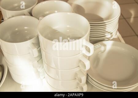 Weiße, keramische, saubere Gerichte in Stapeln, zum Frühstück im Hotel vorbereitet, Gäste erwartet, sonniger Sommermorgen, Sonnenlicht auf Brühe und Teller Stockfoto