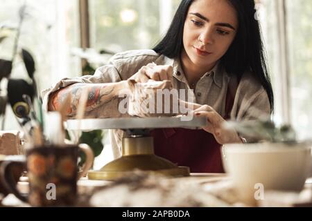 Craftsperson Konzept. Junge Frau, die am Tisch sitzt, um die Keramik im Innenbereich mit dem Modellierwerkzeug zu machen, konzentrierte Nahaufnahme Stockfoto