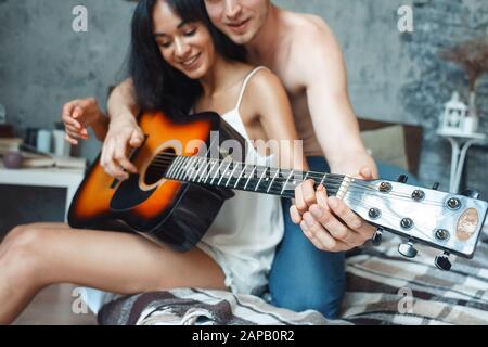 Gemischtes Rennpaar. Junger Mann, der Frau beigebracht hat, Gitarre zu spielen, verschwommener Hintergrund, lächelnd glücklich, zusammen auf dem Bett zu sitzen Stockfoto