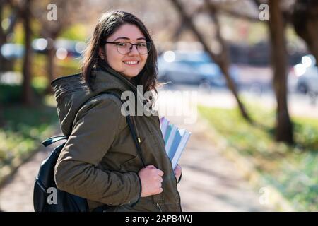 Intellektuelles junges Mädchen bleibt und schaut zu jemandem in einem Park mit einigen Büchern in ihren Händen Stockfoto