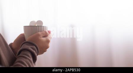 Frau wärmte die Hände und genoss warmes Getränk in der Nähe eines großen Fensters Stockfoto