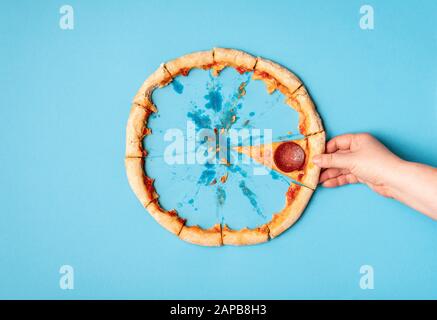 Frau, die ein Stück Pizza Salami und Pizza Kruste übrig hat, auf blauem Hintergrund. Flat Lay vom Essen von Pizza Pfefferoni. Letzte Scheibe Pizza und Kruste. Stockfoto