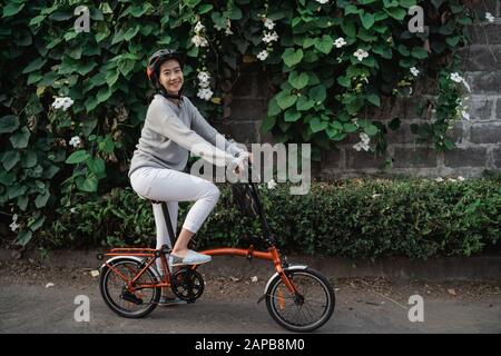 Junge Frauen tragen Helme, um Klappräder im ornamentalen Pflanzenhintergrund zu fahren Stockfoto