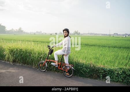 Junge Frauen tragen Helme, um Klappräder im Hintergrund von Reisfeldern zu fahren Stockfoto