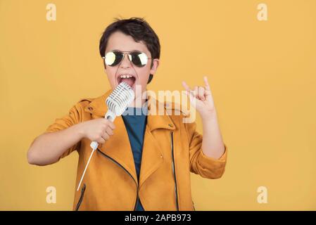 Kind mit Sonnenbrille, das ein Mikrofon hält, das mit den Händen auf gelbem Hintergrund ein Rocksymbol macht Stockfoto