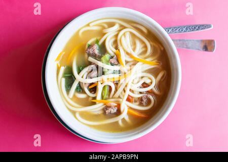 Flacher Lay aus einer Schüssel asiatischer Nudelsuppe, serviert mit Fleisch und Gemüse auf pinkfarbenem Hintergrund Stockfoto