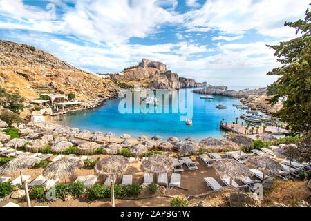 Lindos - Blick auf die Bucht von St. Paul, Strand mit Sonnenliegen und Sonnenschirmen, Akropolis von Lindos im Hintergrund (Rhodos, Griechenland) Stockfoto