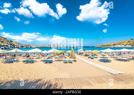 Blick auf den sandigen Strand und die Menschen im Meer in der Bucht von Lindos, Wolken am blauen Himmel (Rhodos, Griechenland) Stockfoto