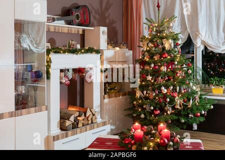 Weihnachten zu Hause im modernen, weißen Wohnzimmer. Der Weihnachtsbaum ist dekoriert. Künstlicher Kamin mit Socken und Weihnachtskranz. Stockfoto